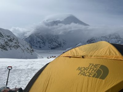 A yellow tent at a snowy mountaineering camp with a cloud-wrapped peak behind it